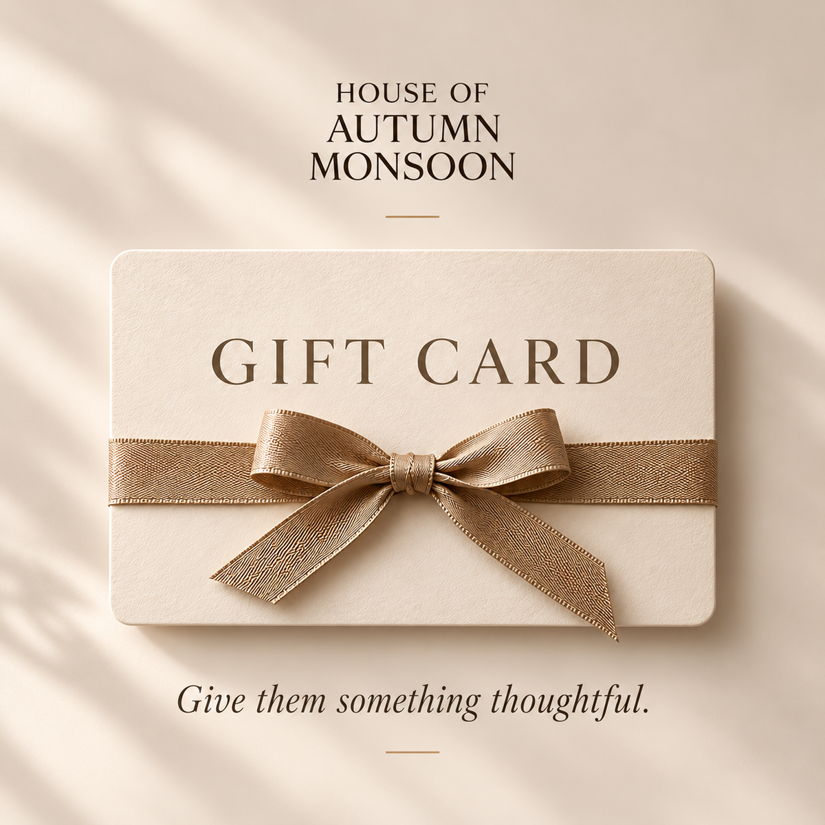 A cream-colored gift card wrapped with a metallic gold ribbon, displayed on a warm beige background with soft leaf shadows. Text above reads 'HOUSE OF AUTUMN MONSOON' and text below reads 'Give them something thoughtful.