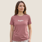 A woman wearing a heathered dusty rose short-sleeve t-shirt with the word 'Inspire' printed in white script across the chest, set against a solid light beige background.