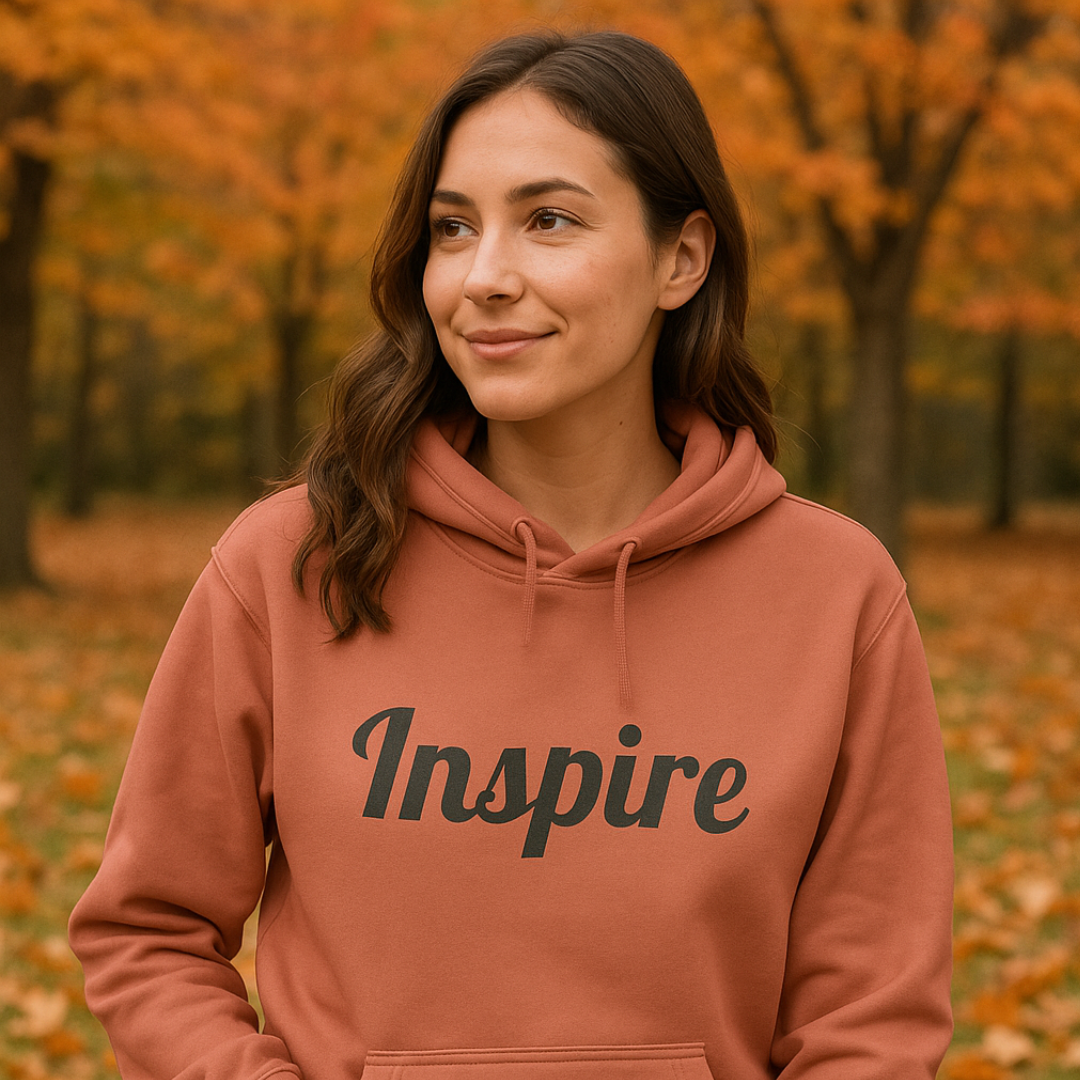 A woman wearing a terracotta-colored hoodie with the word 'Inspire' printed in black script across the chest, standing outdoors in an autumn park with fallen leaves and trees in the background.