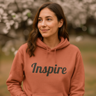 Woman outdoors wearing a dusty rose hoodie that says Inspire.