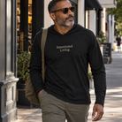 Man walking down a city street wearing a black hooded long sleeve tee with Intentional Living on the chest along with brown pants and carrying a shoulder bag.