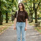 Woman standing on a path in a park wearing a brown t-shirt and light blue jeans.