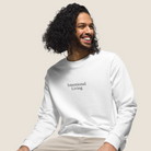 A smiling man with long curly dark hair wearing a white crewneck sweatshirt with the text 'Intentional Living' printed in black on the chest, paired with light beige pants, set against a solid light beige background.