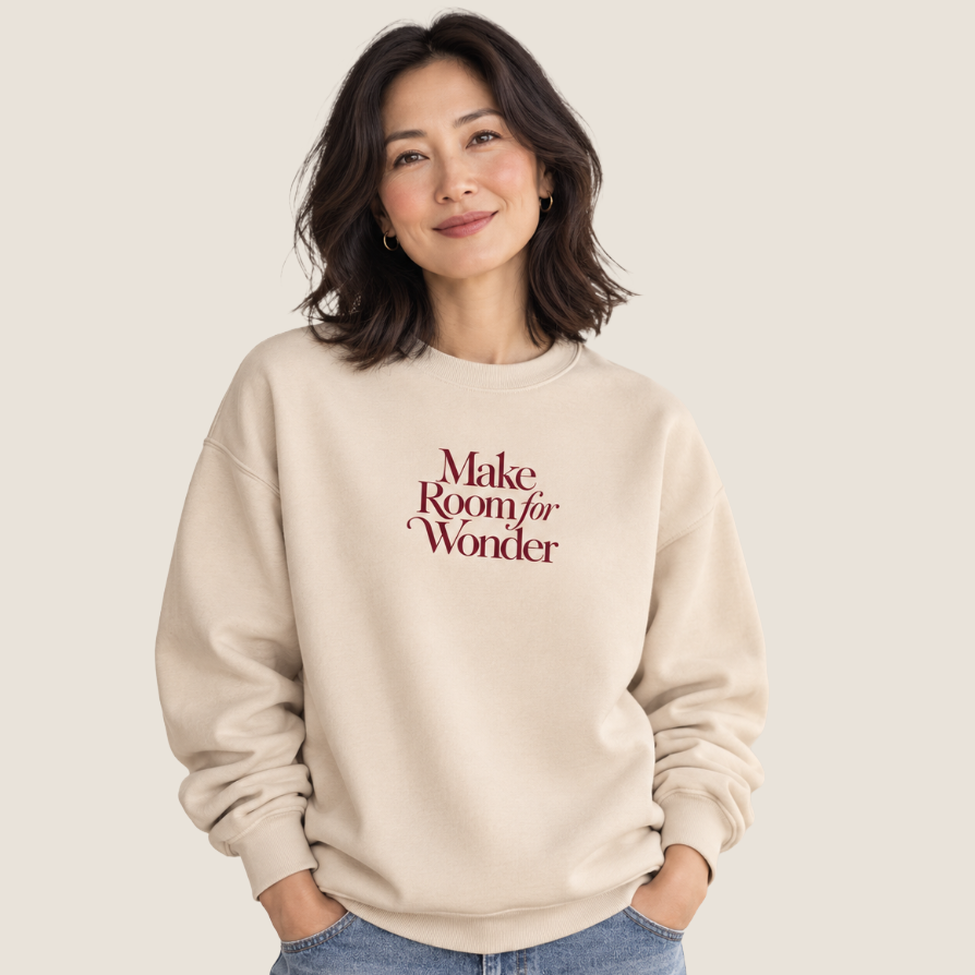 A woman wearing a cream-colored crewneck sweatshirt with the text 'Make Room for Wonder' printed in maroon on the chest, paired with light blue jeans, set against a solid light beige background.