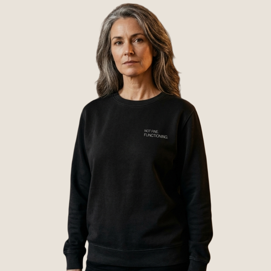 Woman wearing a black Not Fine. Functioning. crewneck sweatshirt against a soft warm beige background. The sweatshirt has a small "Not Fine. Functioning. text at the left chest.