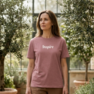 Woman wearing a mauve relaxed tee with the word "Inspire" across the chest. She is standing in a greenhouse.