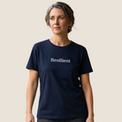A woman wearing a navy blue crewneck t-shirt with the word 'Resilient' printed in white serif text across the chest, standing against a solid light beige background.