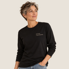 A woman with short gray hair wearing a black long-sleeve t-shirt with the text 'NOT FINE. FUNCTIONING.' printed in white on the left chest, paired with light blue jeans, set against a solid light beige background.