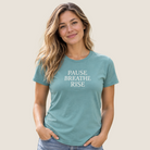 A woman with long wavy blonde hair wearing a sage green graphic t-shirt with the text 'PAUSE BREATHE RISE' in white, paired with blue jeans, standing against a solid beige background.