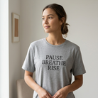Woman wearing a gray t-shirt with 'PAUSE BREATHE RISE' text, standing in a room.