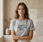 Woman wearing a gray t-shirt with 'PAUSE BREATHE RISE' text in a minimalistic room.