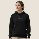 A woman wearing a black pullover hoodie with 'Quiet Motherhood' printed in white text on the chest, paired with beige pants, set against a solid light beige background.
