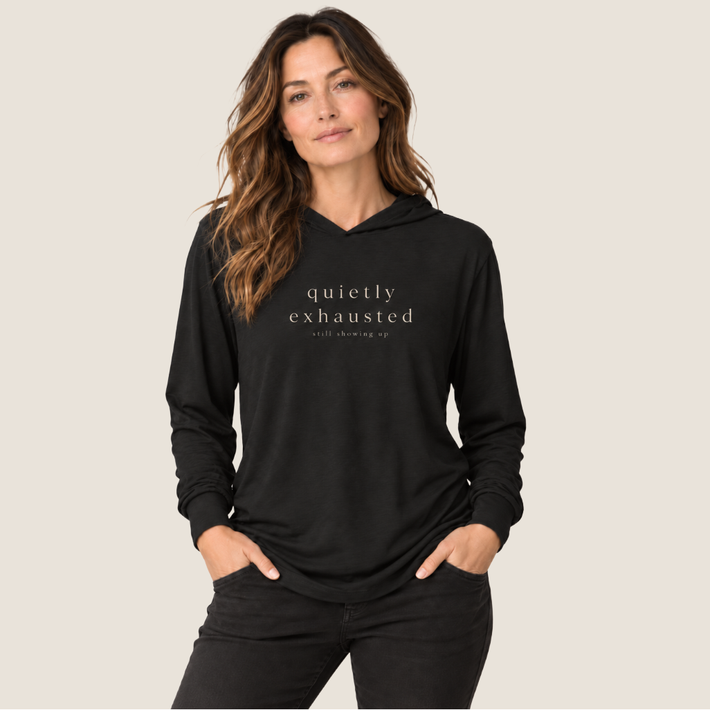 A woman wearing a black pullover hoodie with the text 'quietly exhausted' and 'still showing up' printed in white on the chest, posed against a solid light beige background.