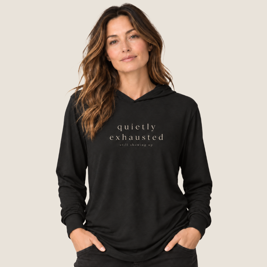 A woman wearing a black pullover hoodie with the text 'quietly exhausted' and 'still showing up' printed in white on the chest, posed against a solid light beige background.