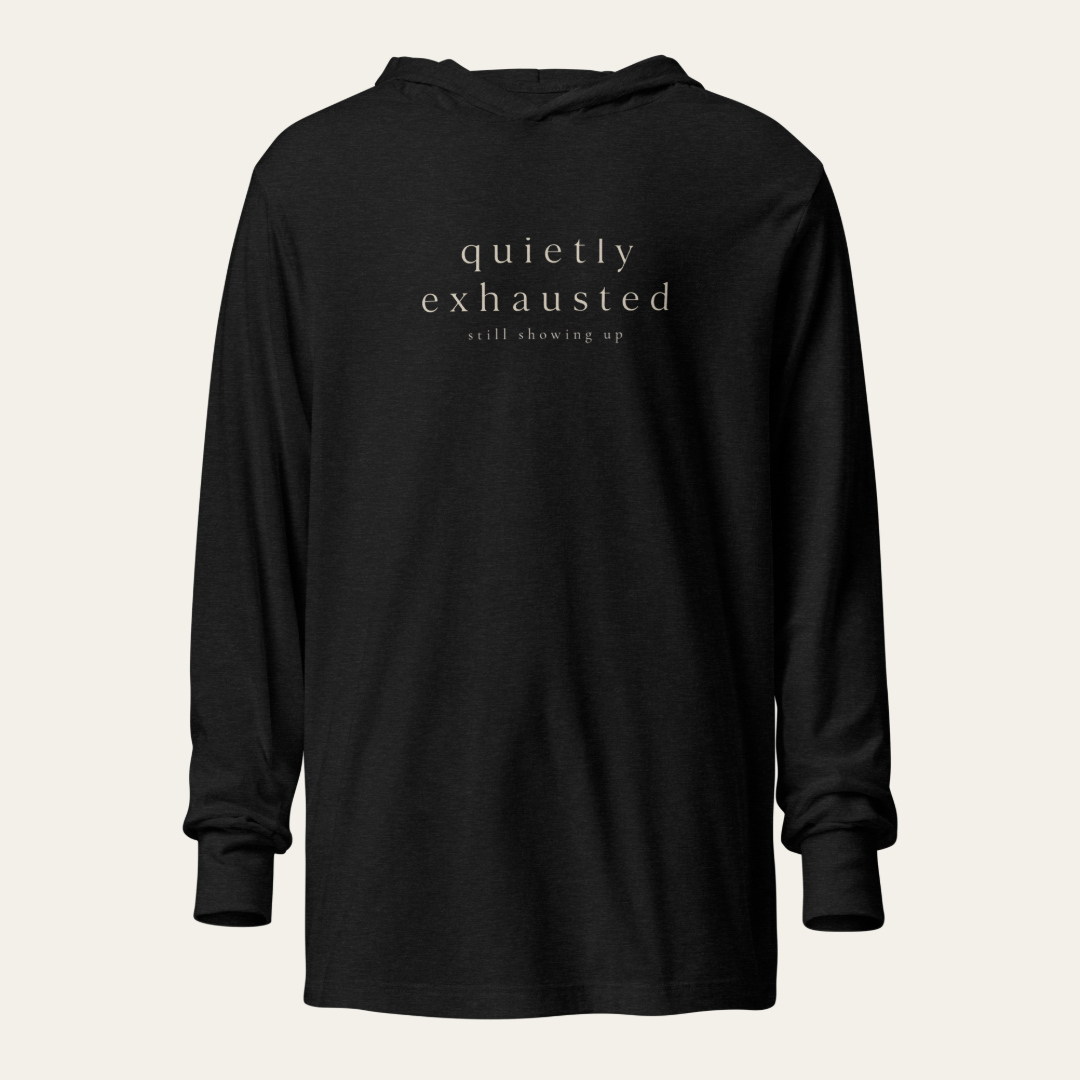 A black long-sleeve hoodie with the words 'quietly exhausted' and 'still showing up' printed in a light beige serif font on the chest, displayed against a light beige background.