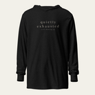 A black long-sleeve hoodie with the words 'quietly exhausted' and 'still showing up' printed in a light beige serif font on the chest, displayed against a light beige background.