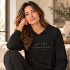 A woman with long wavy brown hair sits on a couch in a softly lit living room, wearing a black long-sleeve hoodie with the words 'quietly exhausted' and smaller text 'still showing up' printed on the front.