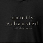 Close-up of a dark charcoal heathered t-shirt with the words 'quietly exhausted' and 'still showing up' printed in a light beige serif font.