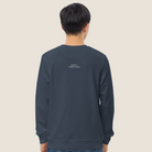 A person viewed from the back wearing a dark slate blue crewneck sweatshirt with small white text on the upper back, paired with blue jeans, against a solid light beige background.