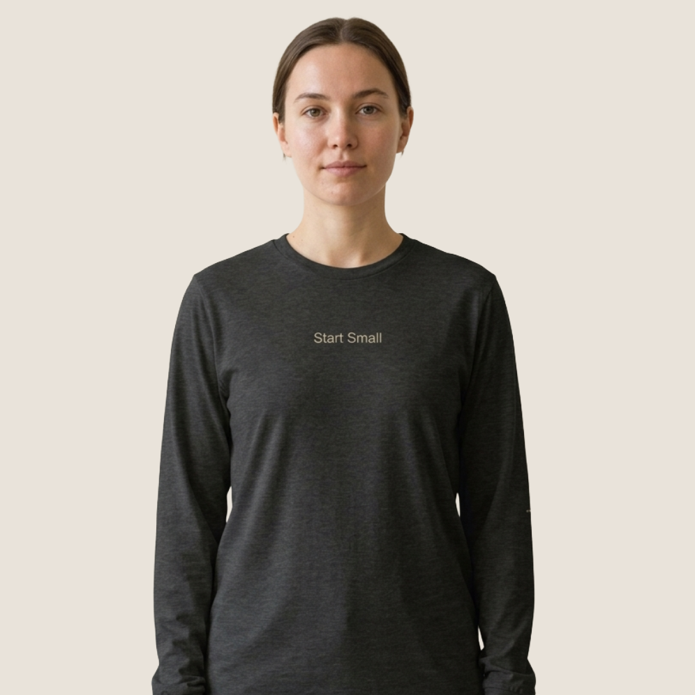 A woman wearing a dark charcoal gray long-sleeve t-shirt with the words 'Start Small' printed in beige on the chest, set against a solid light beige background.