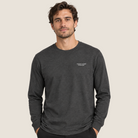 A man wearing a dark heather gray long-sleeve crewneck t-shirt with small white text on the left chest, standing against a solid light beige background.