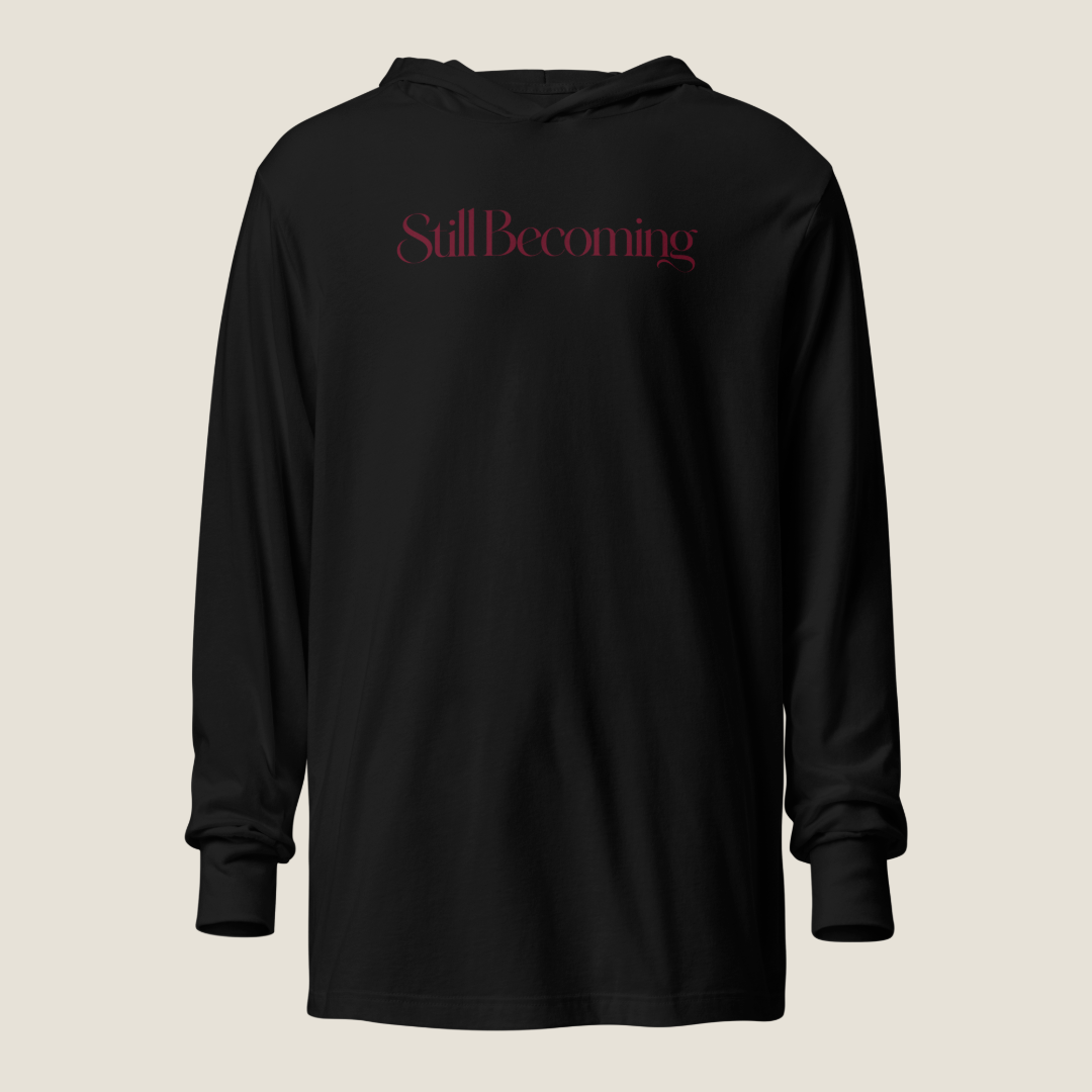 A black long-sleeve hooded sweatshirt with the word 'Still Becoming' printed in a dark red serif font across the chest, displayed against a solid light beige background.