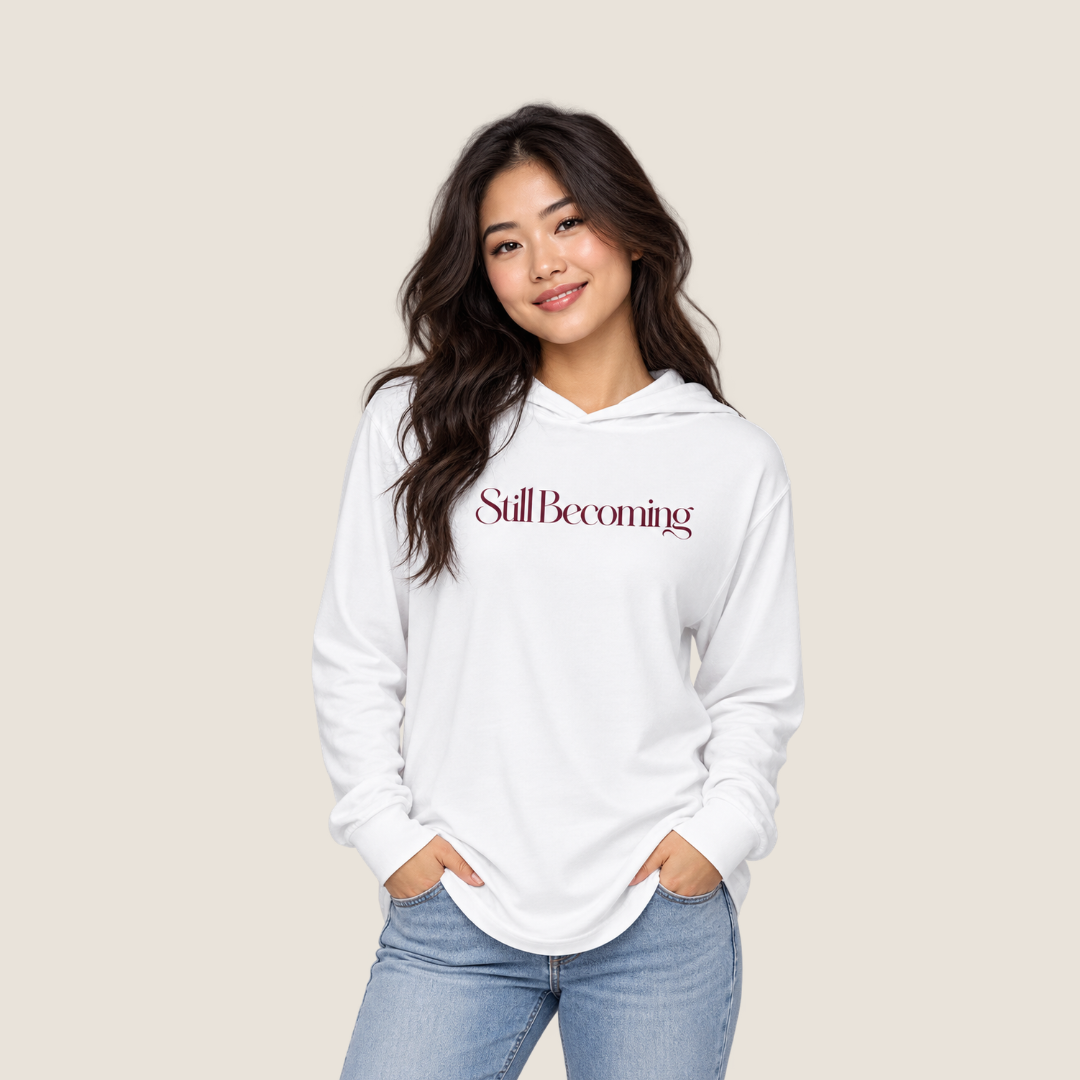 Woman wearing  white hooded long sleeve tee with "Still Becoming" on the chest on a beige background.