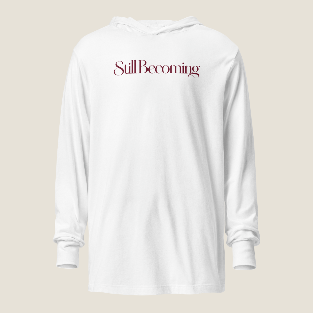 A white long-sleeve hooded sweatshirt with the phrase 'Still Becoming' printed in a dark burgundy serif font across the chest, displayed against a solid light beige background.