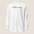 A white long-sleeve hooded sweatshirt with the phrase 'Still Becoming' printed in a dark burgundy serif font across the chest, displayed against a solid light beige background.
