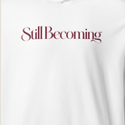 A close-up of a white sweatshirt featuring the phrase 'Still Becoming' printed across the chest in a dark burgundy serif font.