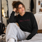 A woman sitting by a bed in a bedroom setting, wearing a black long-sleeve hoodie with the words 'Still Becoming' printed in pink across the chest, paired with light gray sweatpants.