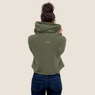 A person viewed from behind wearing an olive green cropped hoodie with small white text on the upper back, paired with dark blue jeans, against a plain light beige background.