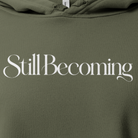 Still Becoming text on an olive green cropped hoodie