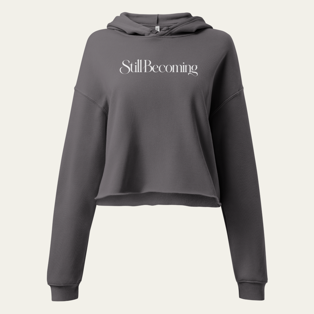A dark gray cropped hoodie with a large graphic of "Still Becoming," displayed on a solid white background.