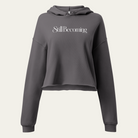A dark gray cropped hoodie with a large graphic of "Still Becoming," displayed on a solid white background.