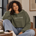 A woman with curly hair sits on a couch wearing an olive green hoodie with the words 'Still Becoming' printed in white across the chest, paired with light blue jeans.