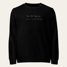 Black crewneck sweatshirt with Still here after everything on upper center chest.