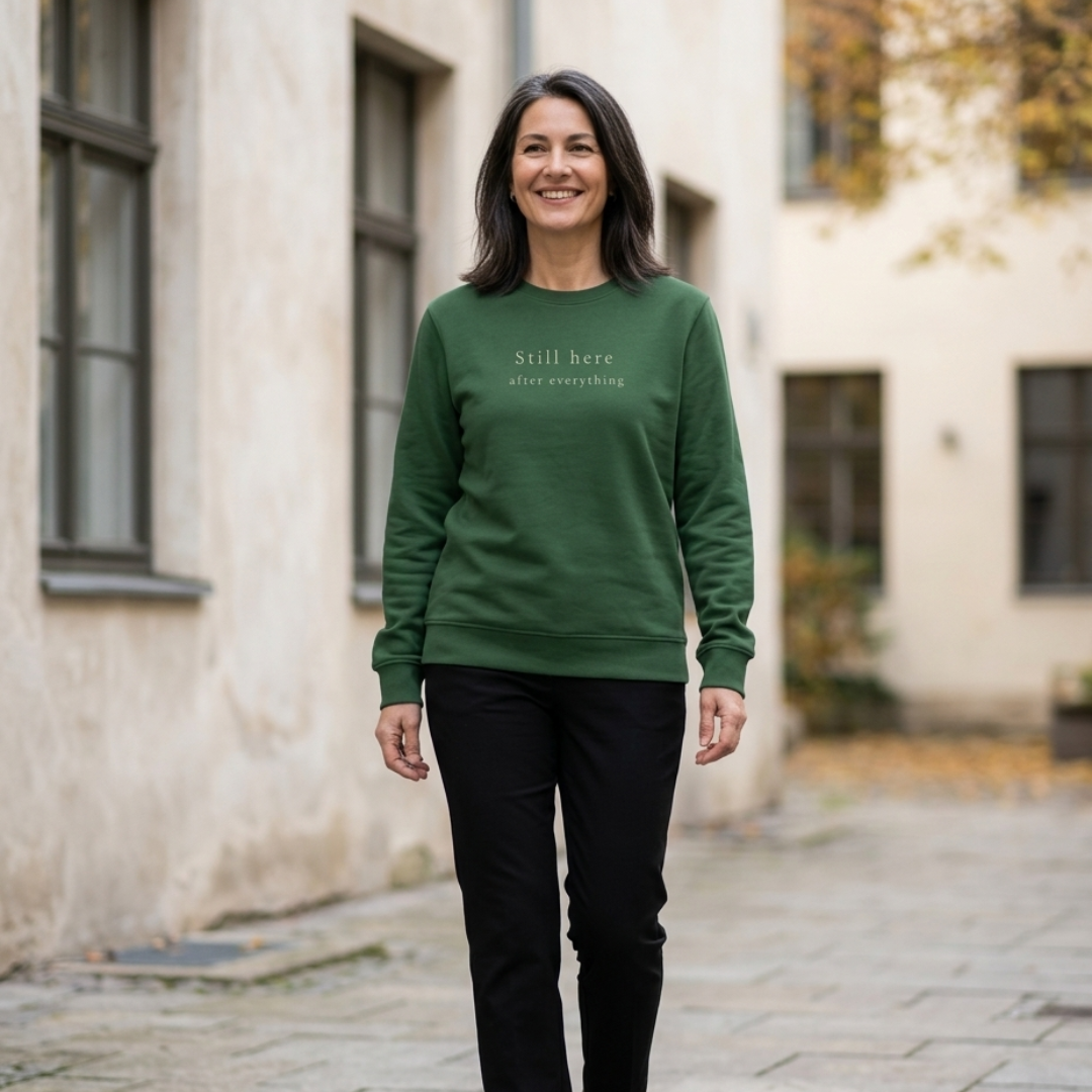 Woman wearing a Still Here crewneck sweatshirt walking down a stone street with stucco buildings in the background.