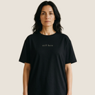 A woman wearing a black short-sleeve crewneck t-shirt with the text 'still here' printed in a small serif font on the chest, set against a solid light beige background.