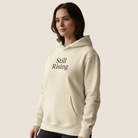 A woman wearing a cream-colored pullover hoodie with the words 'Still Rising' printed in black serif font on the chest, paired with black pants, set against a solid light beige background.