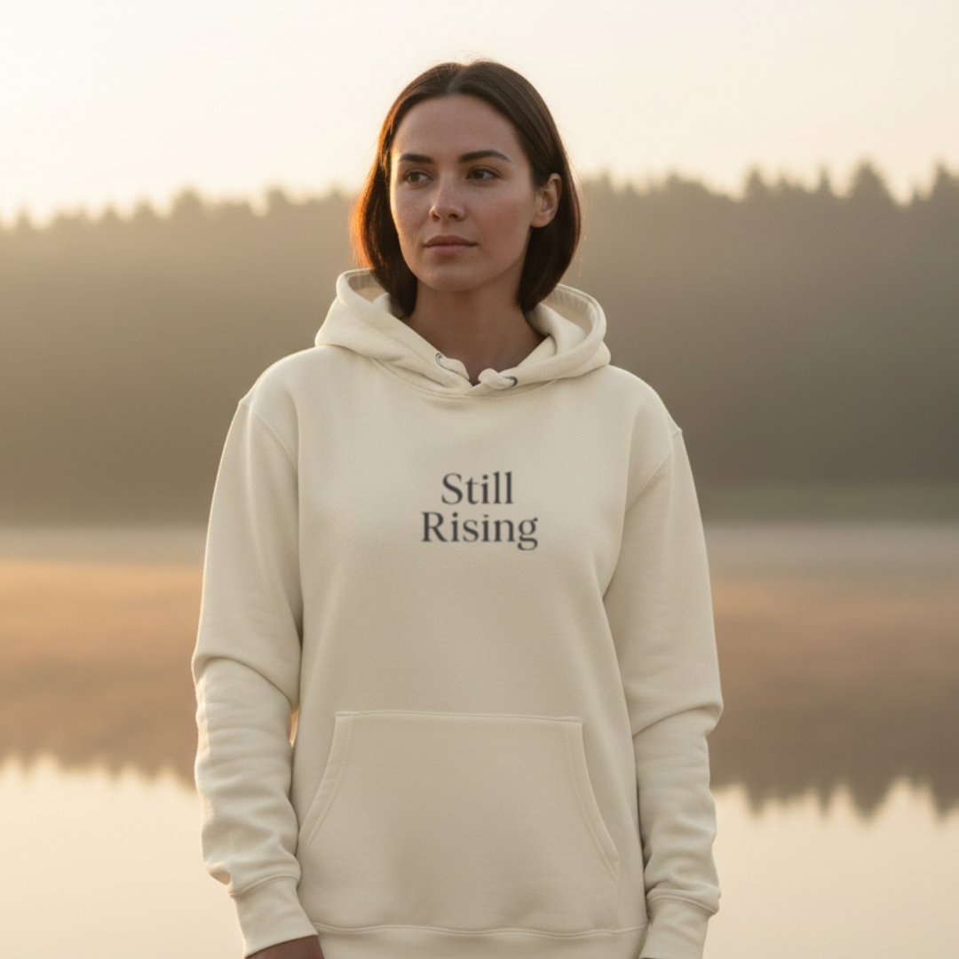 Woman wearing a 'Still Rising' hoodie by a lake at sunset