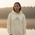 Woman wearing a 'Still Rising' hoodie by a lake at sunset