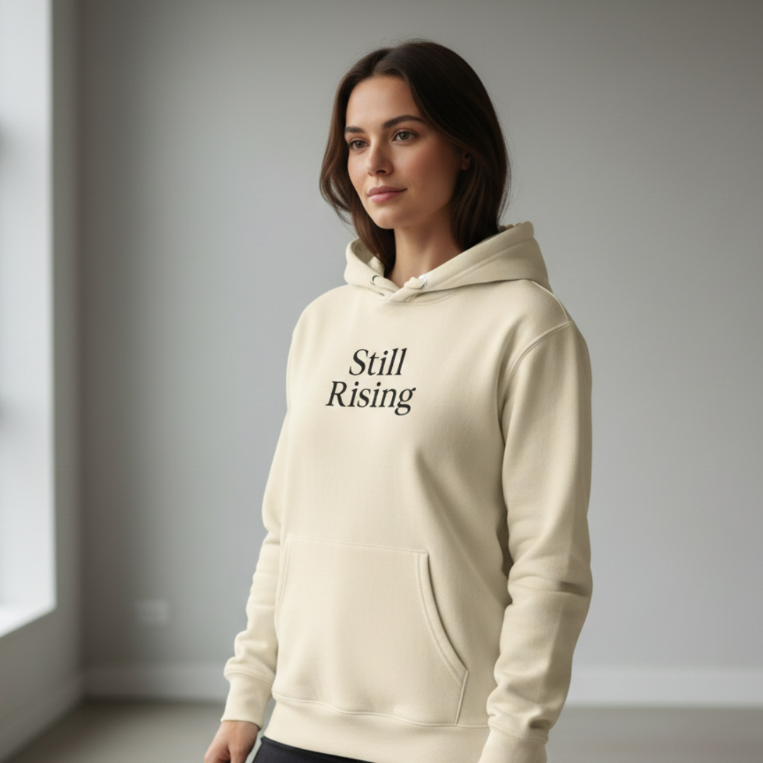 Woman wearing a beige hoodie with 'Still Rising' text in a minimalistic setting