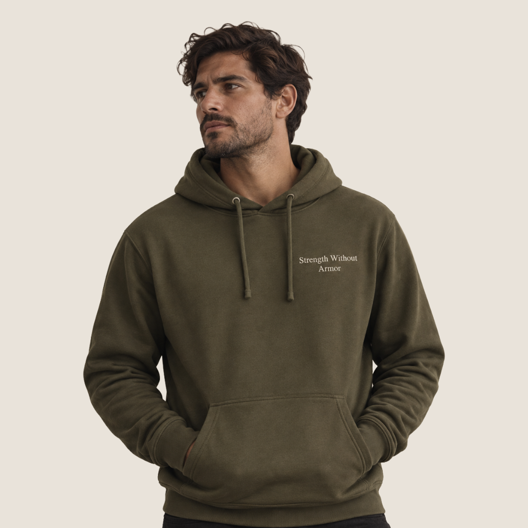 A man wearing an olive green pullover hoodie with the words 'Strength Without Armor' printed in white on the left chest, set against a solid light beige background.