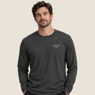 A man wearing a dark heather gray long-sleeve t-shirt with 'Strong in the Quiet' printed in white on the left chest, standing against a solid light beige background.