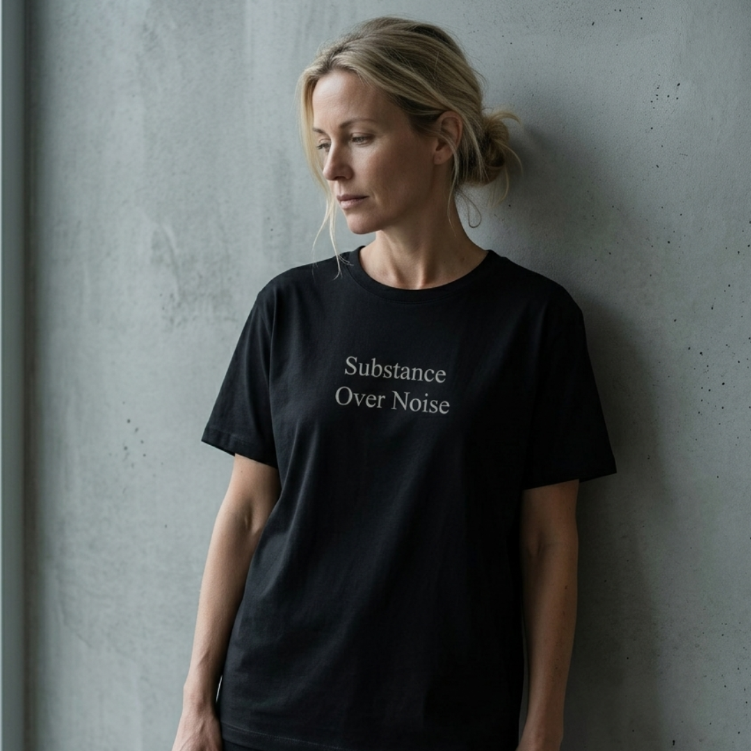 A woman with blonde hair tied back stands against a textured gray concrete wall, wearing a black crewneck t-shirt with the words 'Substance Over Noise' printed in a light beige serif font on the chest.