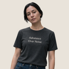 A woman wearing a dark charcoal gray t-shirt with the text 'Substance Over Noise' printed in white serif font, paired with blue jeans, set against a solid light beige background.
