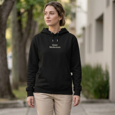 A woman walking outdoors on a sidewalk wearing a black pullover hoodie with 'Quiet Momentum' printed in white on the chest, paired with beige pants. 