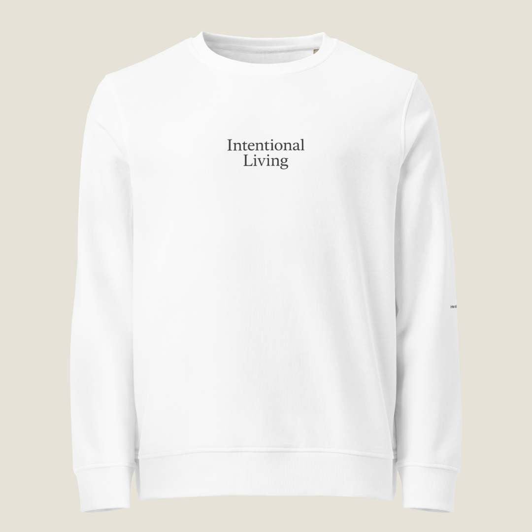A white crewneck sweatshirt with 'Intentional Living' printed in black serif text on the chest, displayed against a solid light beige background.
