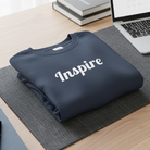 Navy blue sweatshirt with 'Inspire' text on a desk with laptop, coffee, and books.